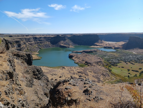 A scenic view of a canyon with a green lake surrounded by rocky cliffs and dry grasslands under a clear blue sky.