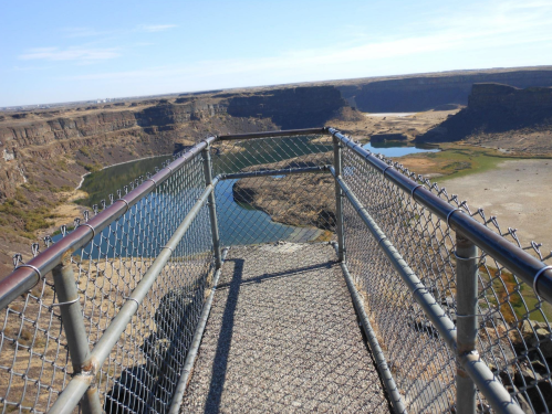 A view from a metal walkway overlooking a canyon with a river and rocky cliffs under a clear blue sky.