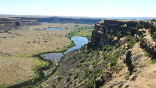 A scenic view of a river winding through a dry landscape, surrounded by cliffs and sparse vegetation under a clear blue sky.