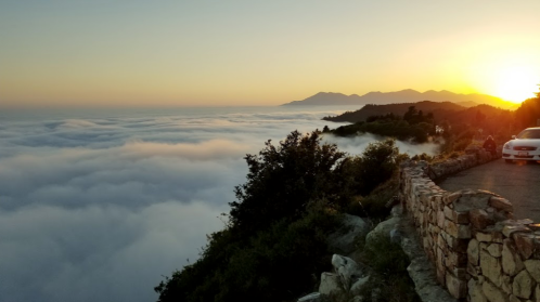 A serene sunset over a sea of clouds, with mountains in the distance and a stone wall along the roadside.
