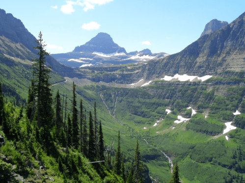 A scenic mountain landscape with lush green valleys, towering peaks, and patches of snow under a clear blue sky.