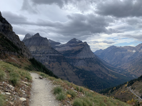 A winding dirt path leads through a mountainous landscape under a cloudy sky, with peaks in the distance.