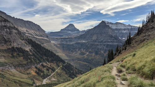 A panoramic view of a mountainous landscape with green hills, rocky peaks, and a winding road below under a cloudy sky.