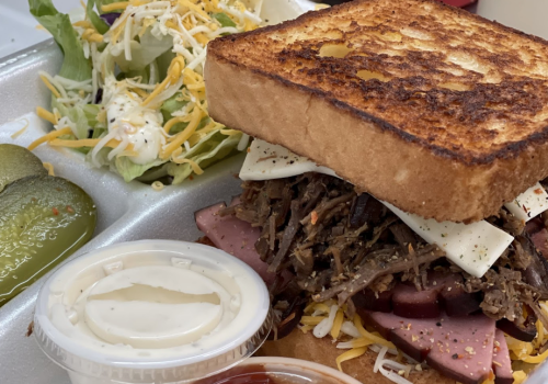 A hearty sandwich with layers of meat and cheese on toasted bread, served with salad, pickles, and dipping sauce.