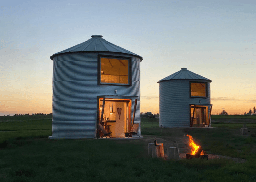 Two converted silos with large windows at sunset, surrounded by grass and a small fire pit in the foreground.