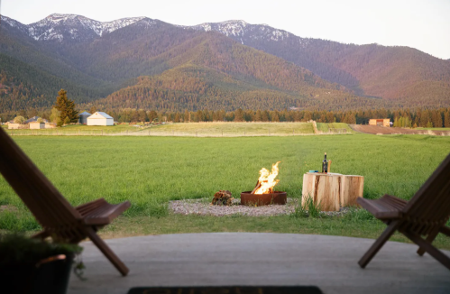 A serene view of mountains and fields from a porch, featuring a fire pit and a bottle of wine on a wooden table.