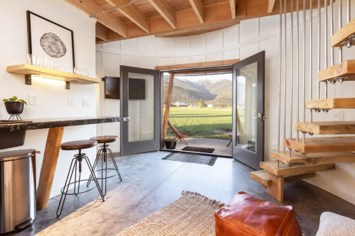 Cozy interior with open doors, modern decor, a staircase, and a view of green fields and mountains outside.