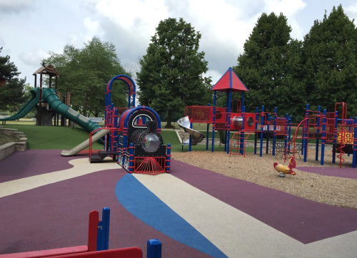 Colorful playground with slides, climbing structures, and a train-themed play area, surrounded by green trees.