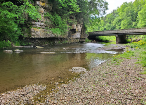 A serene river flows under a stone bridge, surrounded by lush greenery and rocky banks.