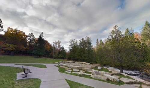 A serene park scene with a bench, winding paths, rocks, and trees beside a flowing river under a cloudy sky.