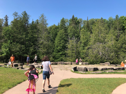 People walking on a path in a park with wooden logs and trees in the background under a clear blue sky.