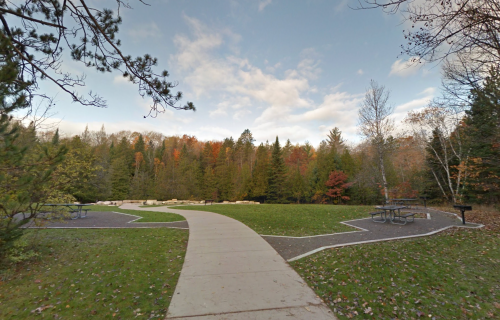 A paved path leads through a park with picnic tables, surrounded by trees displaying autumn colors.