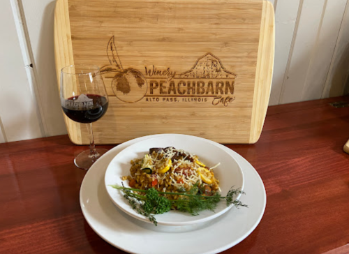 A plate of colorful pasta topped with cheese, alongside a glass of red wine, set against a wooden Peachbarn Cafe sign.