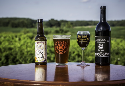 Three beverages on a wooden table: a beer bottle, a glass of beer, and a wine bottle, with a green landscape in the background.