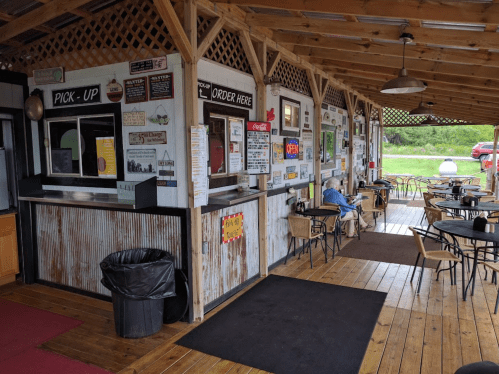 A rustic outdoor dining area with a pickup window, menu boards, and empty tables under a wooden canopy.