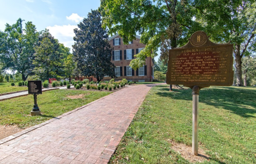 A historic house with a sign, surrounded by greenery and a brick pathway, in a park-like setting.