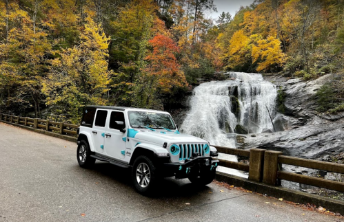 A white Jeep parked by a waterfall, surrounded by colorful autumn foliage.