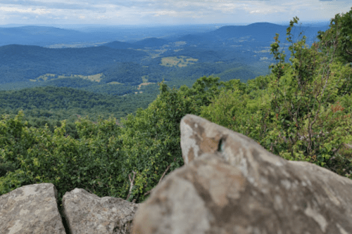 A rocky outcrop in the foreground overlooks a lush green valley and distant mountains under a cloudy sky.
