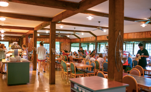 A bustling dining hall with wooden beams, tables filled with people, and staff serving food in a cozy atmosphere.
