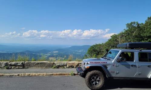 A silver Jeep Rubicon parked at a scenic overlook with a panoramic view of mountains and blue sky.