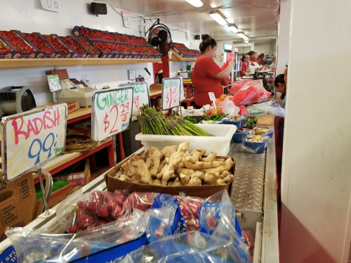 A bustling market stall with fresh produce, including radishes, ginger, and asparagus, with prices displayed.