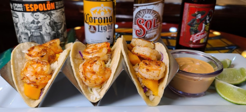 Three shrimp tacos with mango, served with a dipping sauce, lime wedges, and bottles of beer in the background.