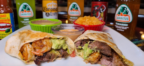 Two burritos filled with shrimp, steak, and guacamole, served with sauces and colorful drinks in the background.