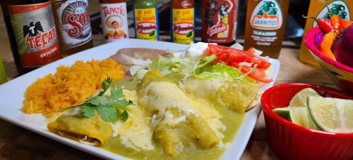 A plate of enchiladas topped with green sauce, served with rice, lettuce, tomatoes, and various hot sauces.