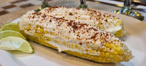 Two ears of corn topped with creamy sauce, cheese, and spices, served with lime wedges on a white plate.