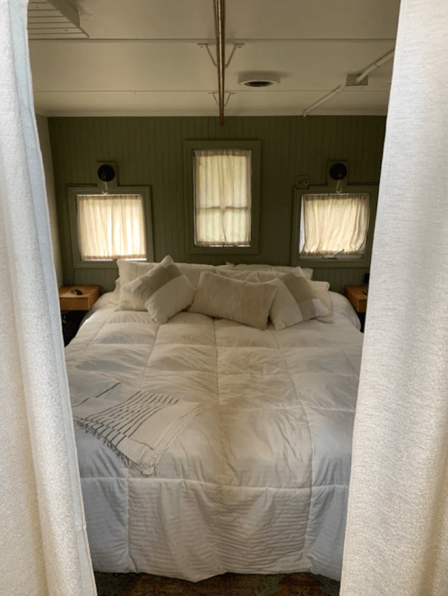 Cozy bedroom with a large bed covered in white bedding, surrounded by soft curtains and natural light from windows.