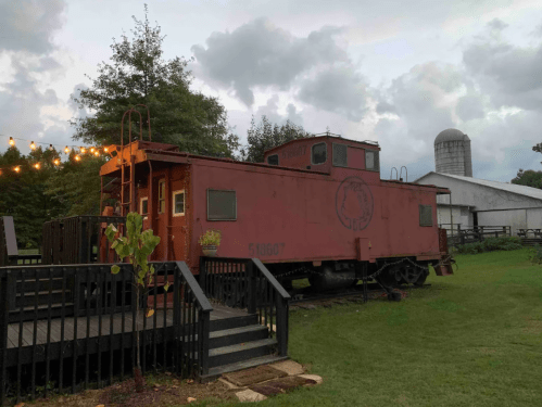 A red caboose on a wooden deck, surrounded by greenery, with a barn and cloudy sky in the background.