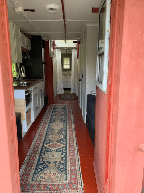 A narrow hallway with a patterned rug, leading to a bedroom, flanked by a kitchen and a cozy seating area.
