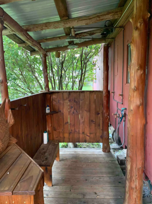 A rustic wooden outdoor shower area with a bench, surrounded by trees and a red wall in the background.