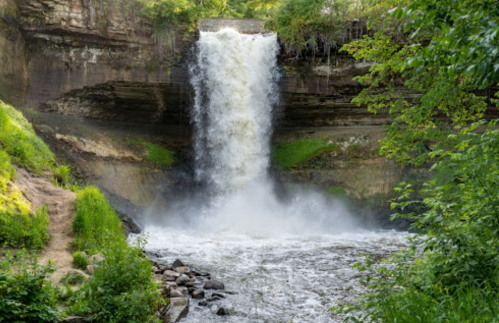 A cascading waterfall flows over a rocky ledge, surrounded by lush greenery and a river below.