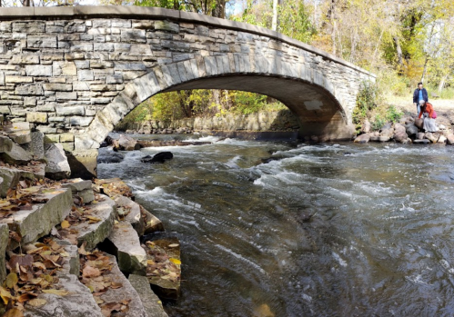 A stone bridge arches over a flowing river, surrounded by trees and autumn leaves on the riverbank.