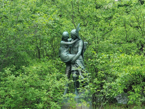 A bronze statue of a person holding a child, surrounded by lush green foliage.