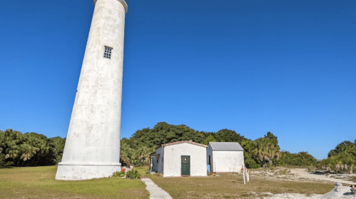 A tall white lighthouse beside a small building, surrounded by greenery under a clear blue sky.