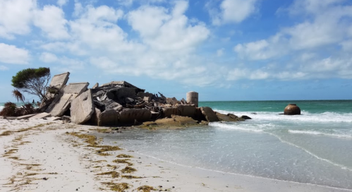A collapsed structure on a beach, with waves lapping at the shore and a cloudy sky above.