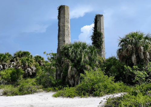 Two weathered concrete pillars surrounded by lush greenery and palm trees on a sandy beach under a blue sky.