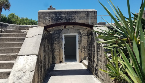 A concrete archway leads to an empty room, surrounded by stone steps and lush greenery under a clear blue sky.
