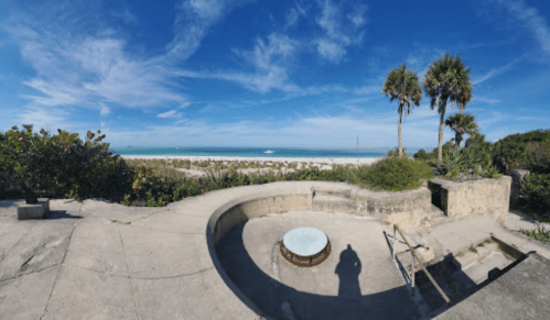 Panoramic view of a beach with palm trees, blue sky, and a circular concrete structure in the foreground.