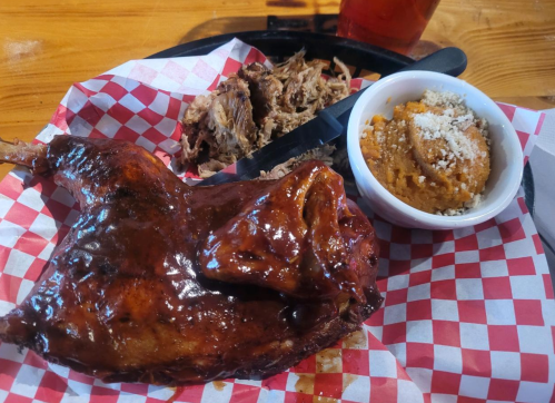 A plate featuring barbecued meat, pulled pork, and a side of sweet potato casserole, served with a drink.