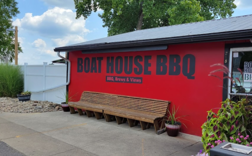 A red building with "BOAT HOUSE BBQ" sign, featuring a wooden bench and greenery in front.