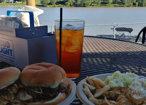 A table with two sandwiches, pasta salad, and iced tea, overlooking a river on a sunny day.