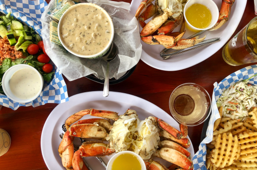 Two plates of seafood featuring crab legs, a creamy soup, coleslaw, and a side salad, with drinks on a wooden table.