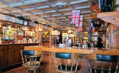 A cozy pub interior with wooden bar, flags, and patrons enjoying drinks. Artwork adorns the walls.