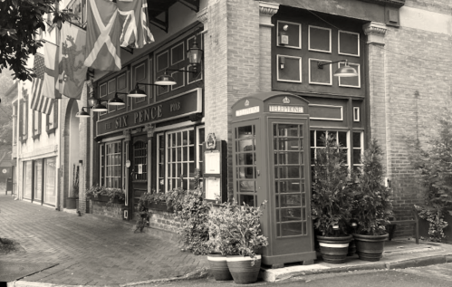 A vintage pub with flags outside, featuring a red telephone booth and potted plants on a cobblestone street.