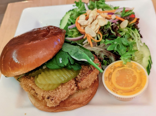 A crispy chicken sandwich with pickles on a bun, served with a side salad and a small container of dressing.