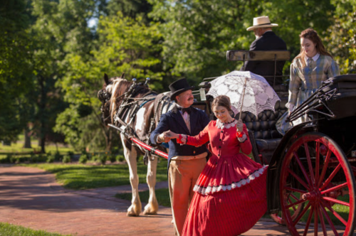 A man in period attire assists a woman in a red dress with a parasol near a horse-drawn carriage in a lush garden.