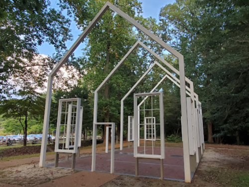 An open-frame structure resembling a house, surrounded by trees, with empty window frames and a clear blue sky.